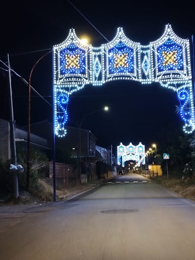 The image depicts a street scene at night in Sessa Aurunca, Italy, illuminated by elaborate decorative arches. In the foreground, a large, ornate archway spans the road, constructed from numerous small LED lights. The primary colors are white and blue, with golden or yellow lights forming geometric patterns, possibly crosses or stars, within square sections of the arch. This style of lighting, known as "luminarie," is characteristic of Italian festivals and religious celebrations.

A paved asphalt road, seemingly empty of vehicles or people, stretches into the distance, reflecting the artificial light. Further down the street, a second, smaller illuminated archway is visible, continuing the festive theme. Streetlights line the road, casting additional illumination.

On either side of the street, residential buildings and dense vegetation are present. Utility poles and overhead wires are visible on the left. On the right side, a blue triangular road sign with a white pedestrian crossing symbol can be seen. The overall impression is one of a quiet town street transformed for a special event, likely a local festa or celebration, common in Italian communities. The time of day is clearly night, under a dark, clear sky. No discernible text beyond the road sign symbol is visible.