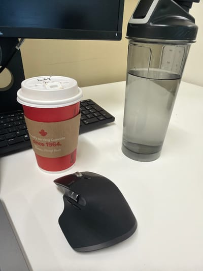 This image depicts a static indoor scene on a white desk, likely within an office setting in Mirabel, Canada. Positioned centrally in the foreground is a red Tim Hortons disposable coffee cup with a white lid. The lid has "LAT" handwritten in black marker. A brown cardboard sleeve around the cup displays a red Canadian maple leaf logo and the text "Proudly serving Canada since 1964. Always Yours." To the right of the cup stands a grey, translucent shaker bottle, approximately half-filled with clear liquid, presumably water, exhibiting visible measurement markings on its side. A black ergonomic computer mouse with multiple buttons and scroll wheels, bearing a "logi" brand inscription, is situated in front of the coffee cup. Partially visible to the left and behind the coffee cup is a black computer keyboard, showing a QWERTY layout with French Canadian keycaps. A dark-colored computer monitor screen and its lower bezel are visible in the background, above the desk, against a plain light beige wall. No individuals are present in the frame, and no actions or direct interactions are occurring.