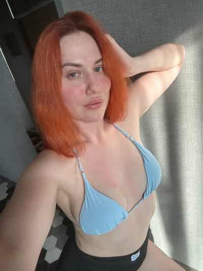 A woman with bright orange hair is taking a selfie in a light blue bikini top and black shorts. Her left arm is raised and touching her hair, while her right arm is down by her side. The background shows a textured gray wall and a black and white tiled floor. There are shadows cast on the wall from what appears to be a window. The location is not visible in the image.