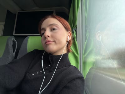 A female subject is depicted seated in a vehicle, likely a bus or train. She has light skin, short reddish-brown hair, and is wearing a black long-sleeved turtleneck sweater with five silver-colored studs/buttons on the left side of the collar. White wired earbuds are inserted into both ears, with the wires visible draped over her chest. She is positioned in a grey and lime-green upholstered seat, with another similar seat visible behind her to the left. An unlit rectangular screen is mounted on the wall above and behind her left. To her right, a window allows bright light, reflecting her profile and a partially drawn pleated green curtain. The environment beyond the window is indistinct due to glare. The specific city location of Courtisols, France, is not visually discernible from the image content.