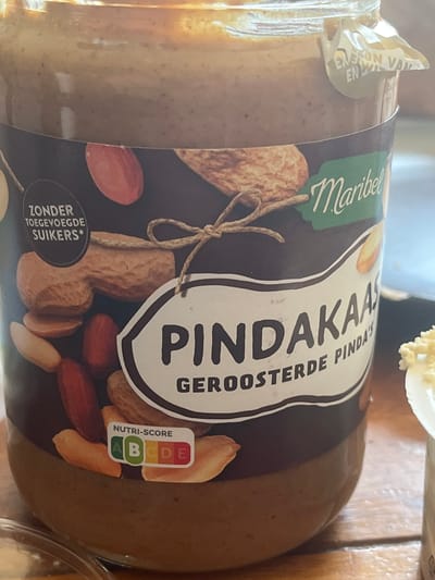 A close-up shot shows a glass jar of Maribel Pindakaas, which is peanut butter. The label on the jar is predominantly dark brown with a large white oval that has "PINDAKAAS" written in large black letters. Below this, "GEROOSTERDE PINDAY" is written in smaller black letters. The word "Maribel" is written in a script font in green above the white oval.
A small black circular label at the bottom left of the jar contains Dutch text that reads "ZONDER TOEGEVOEGDE SUIKERS*", meaning "without added sugars". To the right of this, a Nutri-Score label shows grades A, B, C, D, and E, with "A" and "B" highlighted in green, indicating a relatively healthy score.
Surrounding the central label are images of peanuts, some whole and some shelled, and almonds. A piece of twine is tied in a bow over the peanut images. The jar itself is clear glass, and the peanut butter inside appears smooth and light brown. A small piece of a white lid, possibly holding some other food item, is visible at the top right of the image. The background is blurred but suggests a wooden surface, perhaps a table. The lighting is bright, indicating daytime. There are no people visible in the image.
The overall impression is a product shot focusing on the food item and its nutritional information. The location context provided is Uithuizen, Netherlands, but this information is not directly inferable from the image itself.