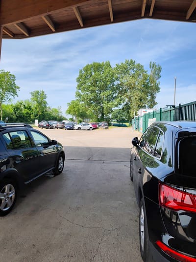 The image shows a view from underneath a wooden shelter or roof structure, likely a parking spot, in what appears to be a parking lot or outdoor area. The location is Leyment, France. A black SUV is parked on the right and a black car is parked on the left, partially obscuring the view into the parking lot. In the parking lot, several other cars of various colors (white, blue, red, dark colors) are parked in rows. Beyond the parking lot, there is a treeline and a fence, and what seems to be a white structure. The sky is visible in the background, with scattered clouds. The ground in the foreground is concrete.
