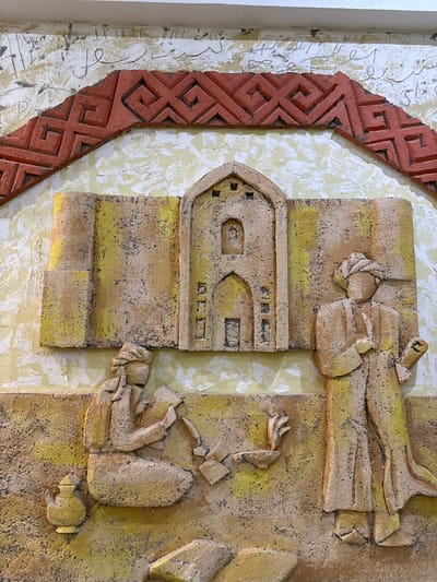 The image displays a bas-relief sculpture depicting a scene with two figures and a building. The artwork is located on a textured wall, with a red, geometric pattern bordering the top of the relief. The central building is rendered in a light brown and yellow hue, featuring an arched doorway and windows. To the left, a seated figure in traditional attire is engaged in an activity, possibly reading or writing, with books and a jug nearby. To the right, a standing figure, also in traditional dress with a turban, holds a rolled document or scroll. The overall color palette consists of earth tones, yellows, and reds, suggesting an arid or historical setting. The sculpture appears to be part of a larger mural or installation in Fort-Şevtşenko, Kazakhstan, likely a cultural or historical monument.