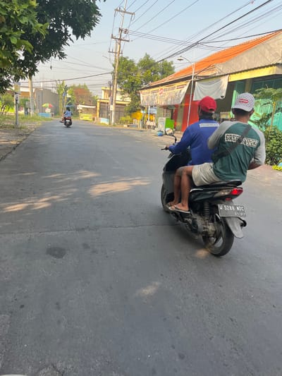 Two men are riding a motorcycle on a paved road in what appears to be an urban or semi-urban area of Karangtanjung, Indonesia. The man in front is wearing a blue long-sleeved shirt with "NORMAT" printed on the back and a red cap. He is driving the motorcycle. The man behind him is wearing a green and gray t-shirt with "GREEN FORCE" printed on it and a white baseball cap with a Supreme logo. He is sitting behind the driver. Both men are wearing shorts and sandals. The motorcycle is dark in color and has a license plate that reads "W 5288 NDC 01-27". The road is asphalt and appears to be in moderate condition with some cracks and dark patches. Sunlight is casting shadows on the road. On either side of the road, there are buildings with orange tiled roofs, storefronts with advertisements, and trees. Power lines are visible overhead. In the background, another person is riding a motorcycle in the distance. The overall scene depicts a typical day on the road in Indonesia.