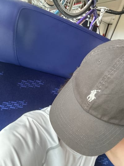 The image shows a close-up view of a person wearing a grey baseball cap with a white polo player logo. The cap is tilted, obscuring most of the person's face. They are wearing a light-colored, possibly white or light grey, athletic shirt. The background reveals a purple bicycle partially visible on the left, suggesting it might be a mode of transport. The person is seated on a blue surface, which appears to be a bench or seat, possibly in a public transportation setting like a train or bus, given the patterned blue fabric behind the person. The overall lighting suggests an indoor environment, possibly during the daytime. The setting appears to be a travel context. No text is clearly visible in the image. The location context provided is Salses-le-Château, France, which could indicate the journey is in or around this area.