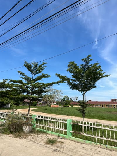 This media file depicts an outdoor scene in Dalam, Indonesia, under a bright blue sky with scattered white clouds, suggesting a sunny day. The image is oriented vertically, with power lines prominently visible in the foreground, stretching diagonally across the frame.

The scene features lush green trees with full foliage, their branches reaching towards the sky. Two larger trees stand out, one towards the top and another further down, both appearing healthy. In the background, a long, low building with a reddish-brown roof and yellow walls is visible, possibly a school or residential complex. A white metal fence with decorative elements runs along the right side of the image, bordering a grassy area.

A dirt path or road curves through the grassy area, leading towards the building. There are no people visible in the frame, and the overall impression is one of a quiet, somewhat rural or suburban environment. The lighting suggests it is daytime, likely late morning or early afternoon. No visible text is present in the image.