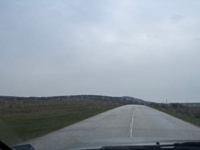 The image depicts a rural road scene, observed from inside a vehicle through a wet windshield, in Gornik, Bulgaria. A damp, two-lane asphalt road with a cracked white center line extends straight into the distance, ascending a gentle incline. Green grassy verges flank both sides of the road, giving way to open fields. The landscape features gently rolling terrain with scattered low-lying scrub brush and a line of hills visible on the horizon. Several utility poles are present in the mid-ground and background, positioned on both sides of the road. The sky is uniformly overcast and grey. There are no individuals, animals, or other vehicles visible in the frame.