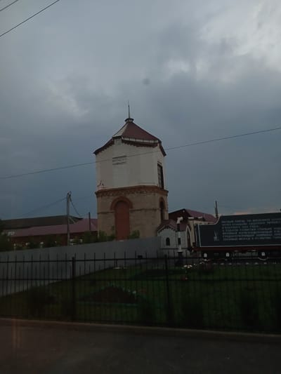 The image depicts a water tower in Abdulino, Russia. The tower is constructed of white brick with a red-tiled roof and a pointed spire. It has a reddish-brown arched doorway on the ground floor. Adjacent to the tower is a dark sign with Cyrillic text, appearing to be a historical marker. The scene is framed by a black metal fence in the foreground, with green grass and some vegetation visible behind it. In the background, other buildings with red roofs can be seen, along with trees and a dark, cloudy sky. The overall impression is of a somewhat overcast day in a semi-industrial or historical district of the city.