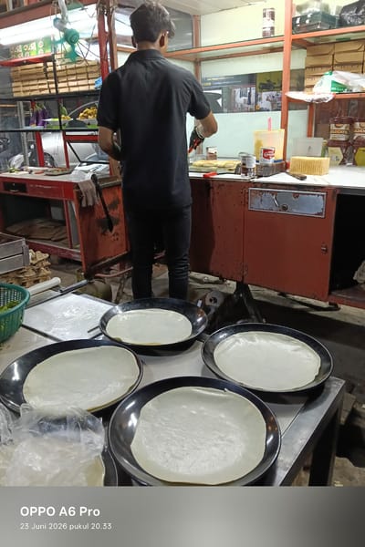This image captures a candid moment at what appears to be a food stall in Sangso, Indonesia. The central subject is a person, likely a vendor, seen from behind, wearing a black polo shirt and dark pants. They are actively engaged in preparing food, holding what looks like a spatula or tongs, and are wearing dark gloves on their hands.
In the foreground, several round, thin, light-colored dough wrappers are laid out on black platters or pans, suggesting they are being prepared for cooking. Some of these wrappers are already in cooking pans, indicating a process in progress. To the left, a green basket contains chopped green vegetables, possibly an ingredient for the food being prepared.
The setting is a busy outdoor or semi-enclosed food stall. Behind the vendor, shelves are stocked with various items, including boxes, containers, and what appear to be canned goods. The lighting suggests it might be evening or nighttime, given the artificial lighting and the general ambiance. Visible text on some of the products includes "Carnation" and "GOLDENFIL," which are brand names for food products. The text "OPPO A6 Pro 23 Juni 2026 pukul 20.33" at the bottom indicates the device and date/time the photo was taken. The overall scene depicts a moment of food preparation and commerce in a local Indonesian setting.