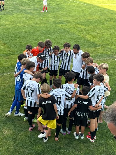 A group of young male soccer players, dressed in black and white striped jerseys, are gathered in a huddle on a green grass soccer field. One player in a bright orange jersey is also part of the huddle. A man in a white shirt and black baseball cap, presumably a coach, is in the center of the huddle, talking to the players. The players are all looking down or towards the coach, with their arms around each other's shoulders. The grass field is well-maintained, and there are faint white lines visible, indicating a soccer pitch. The background shows more of the green field and a hint of a stadium structure in the distance. This scene likely takes place at a soccer stadium in Santa Maria da Feira, Portugal, a city known for its passionate football culture and numerous sports facilities. The players appear to be a team, possibly at a youth league match or training session, and the huddle suggests a moment of team bonding or pre-game preparation.