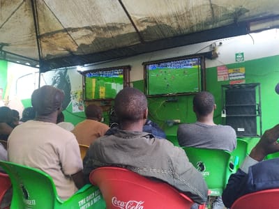 The image shows a group of six men seated in a bar in Sing'isi, Tanzania, watching a football match on two flat-screen televisions. The men are seated at tables, facing the screens, with a large, black speaker in the right background. The bar is adorned with green and white paint, and the men are drinking beer and watching the game. The setting is informal and the men are engrossed in the match.