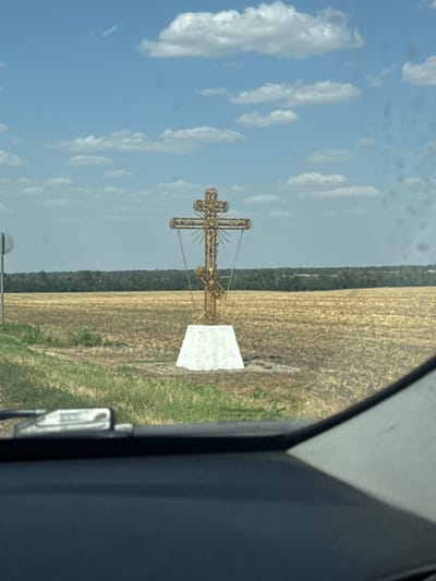 A tall, ornate golden cross stands centered in a harvested wheat field under a bright blue sky with scattered white clouds. The cross is mounted on a white pedestal and appears to be a religious monument. To the left of the cross, a green grassy verge meets the field, and beyond that, a grey pole with a circular sign is visible. The field is dry and golden, suggesting it has been recently harvested. A dense line of dark green trees forms the horizon in the distance. The foreground shows the dark interior of a vehicle, including part of the dashboard and windshield. The image is taken from inside a car, looking out at the landscape. The location is identified as Burdatskii, Russia.