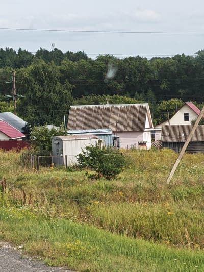 The image depicts a rural scene with several small, weathered houses, likely dacha or summer cottages, nestled among overgrown grass and trees. The main structures include a pink house with a corrugated metal roof, a smaller white building with a similar roof, and another house with a red metal roof in the background. A utilitarian, light-colored metal shed or container is situated in the foreground, with a chain-link fence around it. A thick band of green trees forms the backdrop, suggesting a natural, perhaps forested, environment. The sky is overcast, and a faint trail of smoke or steam rises from a point behind the trees. A utility pole with wires runs across the upper portion of the image. The overall impression is one of a secluded, possibly abandoned or sparsely populated, settlement.