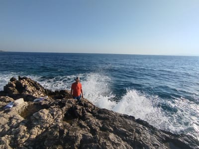 A person stands on the rocky coastline of Karaburun, Turkey, facing the vast expanse of the sea. The individual, wearing a wide-brimmed hat, an orange t-shirt, and blue shorts, is positioned on dark, textured rocks. Waves crash against the shore, sending white spray into the air around the person. The sea is a deep blue, reflecting the clear, pale blue sky. In the distance, the horizon line is sharp, with a faint outline of land visible. There are some white objects, possibly towels or bags, resting on the rocks to the left of the person. The overall scene captures a moment of natural drama and human presence against the backdrop of the Aegean Sea.