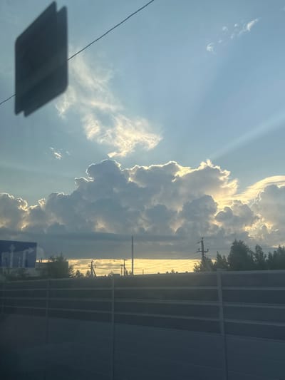 The image captures a sky filled with dramatic, sunlit clouds. The sun's rays are visible, piercing through the clouds and creating a radiant effect. In the foreground, a dark, rectangular object, possibly a sign or part of a structure, is casting a shadow on the sky. Below the sky, there is a landscape that appears to be an urban or semi-urban area, with trees, utility poles, and a large grey barrier or fence. The overall impression is a view of a sunset or sunrise, with a combination of natural and man-made elements. The location is identified as Yarkovo, Russia.