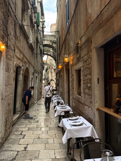 The image depicts a narrow, stone-paved alleyway in Split, Croatia, likely within the historic Diocletian's Palace complex. On the left, a woman wearing a navy blue top and black pants is bent over, looking down at the cobblestones. Further down the alley, a man in a white shirt with dark suspenders and dark pants, presumably a waiter, walks away from the camera. Both sides of the alley are lined with stone buildings, some with arched doorways and windows. The right side of the alley features several outdoor dining tables set with white tablecloths, plates, and silverware, suggesting a restaurant operating in this historic setting. Ornate, amber-lit lamps are mounted on the walls, casting a warm glow. In the background, stone archways span the alley, hinting at the layered architecture of the city. The sky is visible in the distance, appearing overcast.