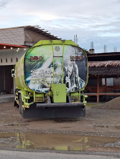 A lime green tanker truck is parked on an unpaved, gravelly surface containing several muddy puddles, under an overcast sky in Keude Lueng Putu, Indonesia. The rear of the truck's tank displays a prominent graphic. On the right, a stylized depiction of Captain Jack Sparrow is featured, with his right index finger held to his lips. To the left, Indonesian text is visible, stating: "Kalau ada Yang ngomongin Dari belakang Berarti kita Lagi di depan" (If someone is talking behind your back, it means we are ahead/in front). This text is accompanied by graphics of lightning bolts, a sailing ship, and a bird. Three circular taillights (one red, two amber) are mounted above the graphic on the tank. A black underride guard is affixed to the lower rear of the truck. A vertical ladder/steps are integrated into the right side of the tank. A circular logo with an intricate design is present at the top center of the tanker. In the left background, a light-colored building with rectangular openings is visible, topped by a building featuring a grey brick facade and a tiled roof with visible rafters. An awning with suspended string lights extends from a structure in the mid-ground. In the right background, a building with a prominent thatched roof and wooden support pillars is present, adjacent to concrete pillars with exposed rebar, indicating construction. A mound of earth or gravel is located in the far right foreground.