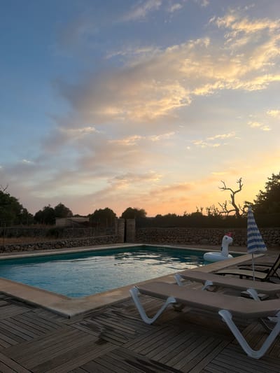 A serene swimming pool area is depicted at dusk in a rural setting. The pool is rectangular with a light blue water surface and a tan concrete border. The surrounding deck is constructed of dark brown wooden planks laid in a diagonal pattern. Two empty, light brown lounge chairs with white frames are positioned on the deck facing the pool. A white inflatable unicorn float with a pink horn is in the pool, and a striped blue and white umbrella stands nearby. In the background, a low stone wall runs along the perimeter, with trees and some simple buildings behind it. The sky is a canvas of soft oranges, pinks, and blues, with scattered clouds, suggesting a peaceful sunset. The location is the city of Santa Margalida, Spain.