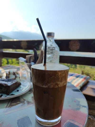 A close-up, eye-level shot captures a frosted glass filled with a dark, frothy beverage, likely a Frappé, with a black straw inserted. The drink is positioned on a patterned, possibly mosaic, table. In the background, a slice of chocolate cake topped with white cream and sprinkled with dark specks sits on a plate to the left. Further back, a textured, clear glass bottle, possibly containing water, stands upright. A dark railing with decorative elements runs horizontally across the mid-ground. Beyond the railing, lush green foliage and distant, hazy mountains under a bright, partly cloudy sky suggest an outdoor setting. The scene appears to be from a cafe or restaurant in Palaios Panteleimonas, Greece, given the traditional architecture often found in such locations and the scenic mountain backdrop. The overall lighting is natural, indicating daytime.