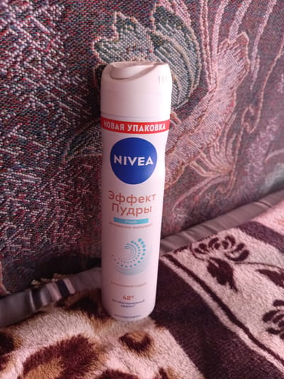 A Nivea "Effect Powder Fresh" aerosol deodorant can stands upright on a textured, possibly woven, surface with a pattern of intertwined green, blue, and reddish-brown threads. The background surface appears to be a blanket or throw. The deodorant can is white with a white cap and a blue circular logo featuring the Nivea name in white letters. Below the logo, text in Russian reads "Эффект Пудры" (Effect Powder) and "Fresh". Further down, it says "Мгновенно высыхает" (Dries instantly). Towards the bottom of the can, there are graphics of small blue circles arranged in a swirling pattern, followed by the text "48ч Антибактериальный эффект" (48h Antibacterial effect) and "Антиперспирант" (Antiperspirant). The overall scene is an indoor shot, and there is no indication of the city of Monastirische, Russia.