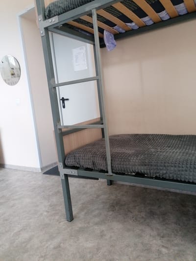 A low-angle, full shot captures a bunk bed in a sparsely furnished room. The frame of the metal bunk bed, painted a muted gray, dominates the foreground and mid-ground. The lower bunk is visible, covered with a textured gray blanket or comforter. The ladder to the upper bunk is also visible, attached to the side of the frame. A white door with a black door handle is partially visible behind the ladder, and a piece of paper is affixed to it. To the left of the bunk bed, a portion of a wall is seen, with a circular mirror mounted on it, reflecting an unseen part of the room. The floor is a textured gray concrete or linoleum. The background wall is a plain, light beige. The lighting appears to be overhead, creating soft shadows. The scene suggests a dormitory or shared living space. The location is identified as Комсомольский, Russia.