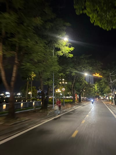 The image depicts a street scene at night in Minh Xuân Ward, Vietnam. The road is wet, suggesting recent rain, and reflects the ambient light from streetlamps and buildings. Lush green trees line the street on both sides, their leaves appearing blurred due to motion. The street is paved and marked with white and yellow lines. On the left side of the road, there is a sidewalk with illuminated rectangular planters or lights. In the background, a tall building is visible with a lit sign at its top. Several streetlights are positioned along the road, illuminating the scene. A few individuals and a motorcycle are present on the street, indicating moderate activity. The overall atmosphere is that of a city street at night, with elements of urban infrastructure and natural foliage.