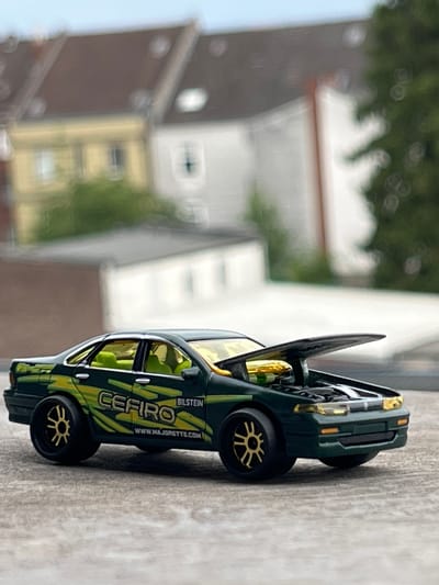 The image shows a close-up, side-view of a green toy car with yellow and black racing stripes. The car is detailed, with visible rims on the wheels, and what appears to be a spoiler on the rear. The car has the word "CEFIRO" printed vertically on its side, along with "WWW.MAJORTOYS.COM" and "BILSTEIN". The toy car is placed on a textured, grey surface, possibly concrete or stone. In the background, out of focus, are buildings and greenery, suggesting an outdoor setting during the daytime. The lighting suggests it is likely daytime with bright conditions. There are no people visible in the image. The overall impression is of a product shot or a display of a collectible toy car, with the background providing context without being the primary focus. The location is Neuss, Germany.