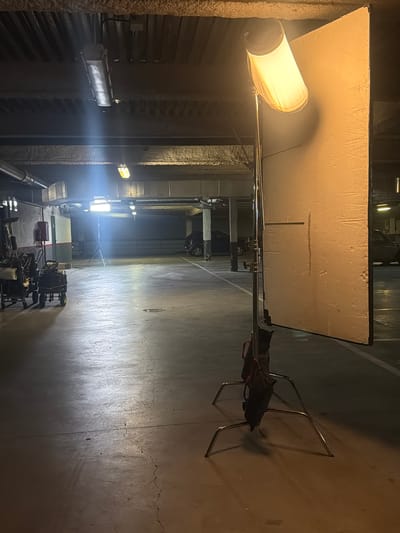This image, captured in San Sebastián De Los Reyes, Spain, displays an artificially lit indoor industrial or studio environment, strongly suggesting a professional production setup for filming or photography.
The scene features a dominant, large, textured, off-white rectangular panel in the foreground, likely serving as a reflector or bounce board. To its left, a cylindrical light fixture with a diffuse cover emits a warm, bright light, casting a distinct shadow on the panel. This light is supported by a metal stand. Another metal stand, carrying a black bag, is visible further to the right.
The concrete floor, showing extensive cracks and wear, is illuminated by various light sources, creating bright reflections. In the midground, several powerful, cool white light fixtures cast strong beams across the space, highlighting the floor's surface and the industrial structures along the walls, which appear to be rough concrete or insulated panels with horizontal elements.
In the background, a collection of equipment on wheels, possibly including camera rigs, generators, or lighting control units, is arranged on the floor, alongside a red toolbox. A dark vehicle is partially visible further in the distance, indicating a large, accessible space. There are no people present in the image, and no visible text. The overall ambiance is functional and focused on the technical aspects of a production, with a clear emphasis on illumination and equipment arrangement.