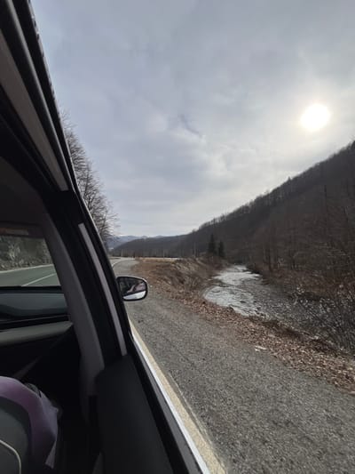 The image provides a view from the passenger seat of a vehicle in motion through a mountainous region in Lugovi, Montenegro. The car's black A-pillar and part of the passenger door frame are visible on the left. Through the windows, a two-lane paved road with a solid white fog line and a solid yellow center line is visible, curving to the left in the distance. To the immediate right of the road is an unpaved shoulder composed of gravel and brown, sparse vegetation. Further right, a flowing river or stream with a rocky bed runs parallel to the road; its banks display some brown foliage and potential ice patches. The landscape is dominated by steep hillsides and mountains covered predominantly with barren deciduous trees, indicating a cold season, possibly winter or early spring. A few coniferous trees are interspersed near the river. The sky above is partially cloudy, with a bright, intense sun visible in the upper right quadrant of the frame. A blurred reflection in the car's side mirror shows an indeterminate light-colored object or figure.