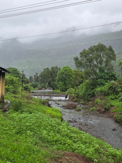 The image depicts a cloudy, overcast day in the rural outskirts of Sandshi, India. A small, concrete dam with water flowing over it bisects a rocky stream in the foreground. Lush green vegetation blankets the landscape, with dense trees and shrubs lining the banks of the stream. In the background, mist-shrouded hills rise, with faint waterfalls visible cascading down their slopes. To the left, a portion of a yellow-colored building with a dark roof is visible, possibly a small dwelling or roadside structure. A dirt path is discernible in the lower portion of the image, leading towards the stream. Overhead, power lines stretch across the frame, indicating a rural, yet connected, area. A yellow vehicle and a few people are visible near the dam in the middle ground, suggesting minimal human activity in this natural setting. The overall impression is one of serene natural beauty, characteristic of the monsoon season in a hilly region.