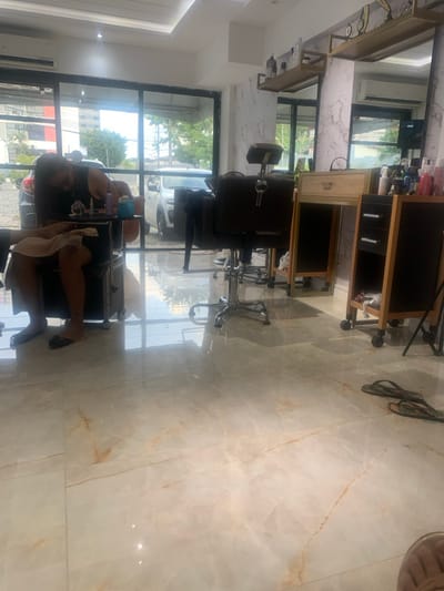 The image depicts the interior of a beauty salon or similar establishment in Lauro de Freitas, Brazil. The flooring consists of large, polished, light-colored tiles with subtle brown and orange veining, creating a reflective surface.
On the left side of the frame, a person with long dark hair, wearing a dark sleeveless top and a light-colored skirt, is seated in a dark salon chair, facing away from the camera. Their hands are positioned over a small, dark mobile trolley in front of them, which holds several small bottles, tools, and a blue cup or bowl, indicating the performance of a nail service. A light-colored towel is draped over their lap and legs. A small dark tattoo is visible on the person's left ankle.
To the right of the seated individual, an empty dark salon chair with a chrome base is positioned. Further to the right, against a white wall with a marble-like pattern, two mobile wooden trolleys are present. The first is a light wood color with a single drawer and a mirror above it, topped by a gold-colored metal shelf displaying decorative items. The second trolley, to its right, is a darker wood with multiple shelves, holding numerous cosmetic bottles and products. Overhead, the ceiling is white with recessed lighting.
Through the large glass windows comprising the left and center background, an outdoor parking area is visible. A dark grey SUV is parked directly outside the windows on the left, and a white sedan is parked to its right. Beyond the vehicles, green foliage and trees are visible, with distant buildings under a clear sky, including a multi-story structure with red and white sections. A pair of dark, patterned flip-flops are on the tiled floor in the lower right portion of the image.