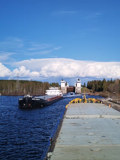The image depicts a canal scene in Анхимово, Russia, showcasing a barge named "ВОЛГО-ДОН 101" transiting through a lock. The sky is partly cloudy with a prominent bank of cumulus clouds. In the foreground is a flat-deck barge with yellow machinery, and in the distance, the lock structure is visible. The surrounding environment includes forested areas and an electrical transmission tower. The overall scene suggests a waterway used for industrial or commercial transportation.