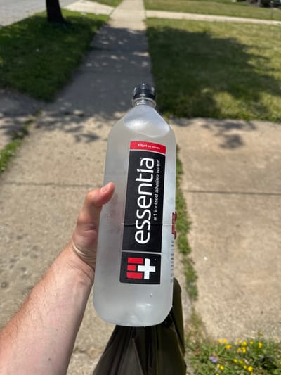 The image shows a person's hand holding a bottle of Essentia ionized alkaline water. The bottle is clear and filled with water, with condensation on the outside. The label on the bottle is black and white, with the Essentia logo and the text "#1 ionized alkaline water" and "9.5pH or higher". The hand holding the bottle is a light skin tone with some arm hair visible.
The scene appears to be outdoors, with a sidewalk and grass visible in the background. There are shadows cast on the ground, suggesting it is daytime, likely with bright sunlight. In the distance, some yellow wildflowers can be seen. The overall impression is of a casual, everyday moment, possibly a walk or a break outdoors. The focus of the image is clearly on the bottle of water and the hand holding it. No specific emotions are conveyed, but the setting suggests a relaxed atmosphere. The location is identified as Maple Heights, United States of America.