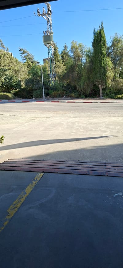 The image shows an outdoor scene with a clear blue sky. In the background, a tall electricity pylon with a transformer is visible, surrounded by trees. A street runs horizontally across the middle of the image, with a red and white striped curb. In the foreground, there is a concrete surface with a metal speed bump and a faded yellow line painted on the ground. The scene appears to be in a semi-urban or suburban area, possibly near a road, with vegetation and infrastructure present. The specific location in the Jezreel Valley Regional Council, Israel, is not explicitly depicted by any landmarks or signs within the frame.