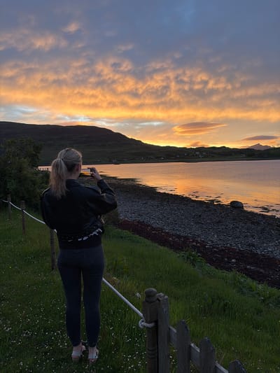 An outdoor scene captures a person observing and photographing a sunset over a body of water in Sconser, United Kingdom. A blonde individual, seen from behind, is centered in the midground, wearing a black leather jacket, dark trousers, and light-colored sandals. Their hair is tied in a ponytail. They hold a smartphone horizontally at eye level, actively capturing the vivid orange and gold sunset reflected across the calm water.

The sky above displays dramatic orange, gold, and light blue clouds, indicating late evening. Dark, rolling hills and mountains frame the distant horizon, with scattered small buildings visible on the far side of the water, nestled at the base of the hills. The water's surface reflects the warm hues of the sky. The immediate foreground features a green grassy slope with sparse white wildflowers, delimited by a wooden fence with a white rope railing that extends into the scene from the lower right and left. Beyond the grass and fence, a rocky, pebbled shoreline with patches of dark seaweed transitions into the water. A single larger rock rests on the pebbled shore to the right.
