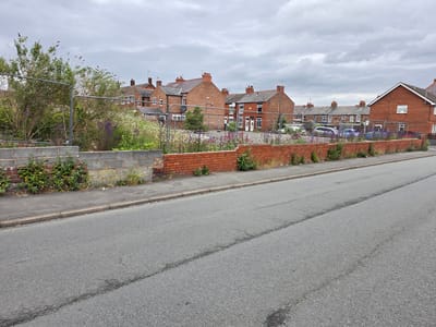 This image depicts a residential street scene in Shotton, United Kingdom, under an overcast sky. In the foreground, an asphalt road runs diagonally from the lower left to the upper right, exhibiting a visible crack or seam across its width. A concrete curb separates the road from a paved sidewalk, which runs along the bottom left edge of the frame.
Adjacent to the sidewalk, a low retaining wall is present. The left portion of this wall consists of stacked concrete blocks, partially obscured by green vegetation growing both against it and from its top. The right portion of the wall is constructed from red bricks, also showing some sparse green and dry vegetation growing along its base and top.
Behind this wall lies an overgrown plot of land, currently secured by galvanized metal temporary fencing panels that span horizontally across the middle ground of the image. This plot is densely covered with various plants, including tall green bushes with dark seed heads or small flowers (potentially Buddleia), numerous white wildflowers resembling daisies, and clusters of distinct purple flowers.
In the background, beyond the fenced plot, a row of two-story red-brick residential houses is visible. These houses feature pitched roofs, several prominent chimneys, and multiple television aerials on their rooftops. The architectural style suggests typical terraced or semi-detached British housing. Some parked cars are discernible through the gaps in the vegetation and fencing within the plot, positioned closer to these houses. No individuals or active interactions are visible within the frame.