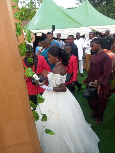 In Amokvo, Nigeria, an outdoor celebratory event is captured, featuring a woman in bridal attire and multiple attendees. The central figure is a woman of dark complexion, wearing a white off-shoulder wedding gown embellished with beading, a pearl necklace, and white lace fingerless gloves. She stands facing left, her hands occupied with small objects, indicative of a transaction or gift exchange. To her left, partially obscured, a man of dark complexion in a red suit jacket with black lapels also handles small items. To the bride's right, a man of dark complexion with facial hair wears a maroon traditional Nigerian garment and holds a black plastic bag and a brown object, possibly a document or folder, in his left hand.
Numerous other individuals, predominantly men in various forms of dress including traditional Nigerian wear and Western attire, populate the background. Some are actively observing, while one individual in the mid-background holds a smartphone aloft, potentially recording the event. The scene takes place on green artificial turf under large light-green canvas event tents, which are draped with white fabric underneath. A tall, light-brown vertical structure adorned with artificial green ivy vines is prominent on the far left. Trees and foliage are visible above the tents, indicating an open-air setting.