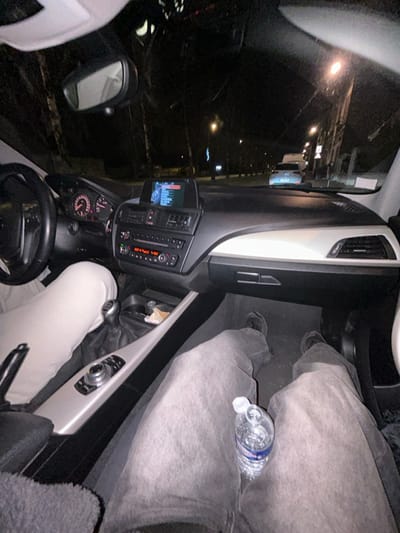 Inside a car at night, the driver's perspective is seen. The steering wheel, dashboard with a screen and radio, and gear stick are visible. The driver wears white pants, while the passenger's side shows gray pants with a water bottle nearby. Outside the window, streetlights illuminate buildings and other cars parked along the street.