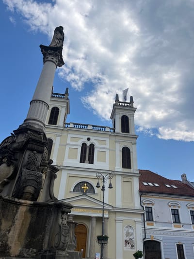 This is a low-angle, outdoor, daytime, full-shot photograph taken from the front of the Church of the Assumption of the Virgin Mary in Banská Bystrica, Slovakia. In the foreground on the left is a stone column topped by a statue of a person. The column has intricate carvings at its base, and a lamp post with multiple lights stands to the right of it. Behind the column, the pale yellow facade of the church is visible. The church has two square towers with dark shutters on the windows and is topped with a flag on the right tower. A gold cross is mounted above the entrance to the church, which has a large wooden door. To the right of the church, a reddish-tiled roof of a building is visible. The sky is blue with scattered white clouds.