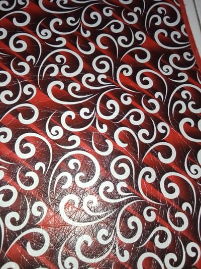 The image displays a close-up view of a patterned surface, located in Ciputri, Indonesia. The surface features a primary design of broad, diagonal stripes in varying shades of red and dark reddish-brown, running from upper-left to lower-right. Superimposed over these stripes is a dense, repeating pattern of white, curvilinear, scroll-like motifs that vary in size and orientation. The material exhibits a slight sheen and numerous fine, intersecting lines or textures are visible, particularly within the darker red areas, indicating a textured or possibly worn finish. No individuals, discernible objects other than the surface itself, or actions are depicted. The environment is limited to this patterned material.