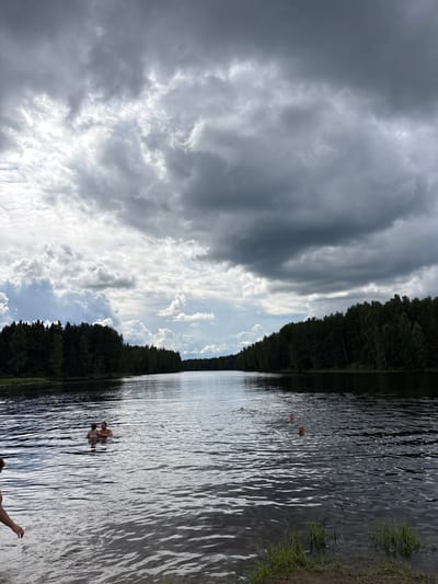 This image depicts a wide, calm lake with dark, rippling water. On the left side of the foreground, a person's arm and shoulder are visible, emerging from the water. Further into the lake, a couple stands waist-deep, facing each other. In the mid-distance, three other individuals are swimming, with only their heads visible above the water. The lake is bordered by a dense forest on both sides, with tall, dark green trees lining the shore. The sky above is overcast with dramatic, grey storm clouds, interspersed with patches of brighter sky. The overall scene suggests a natural, outdoor environment, likely a recreational area. The location is identified as Burtsevo, Russia.