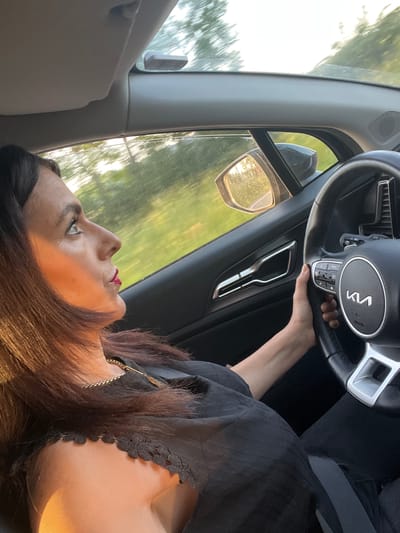 A woman is in the driver's seat of a car. The car has a Kia steering wheel. The side mirror is visible. The environment outside the car appears to be trees. The location is unknown.