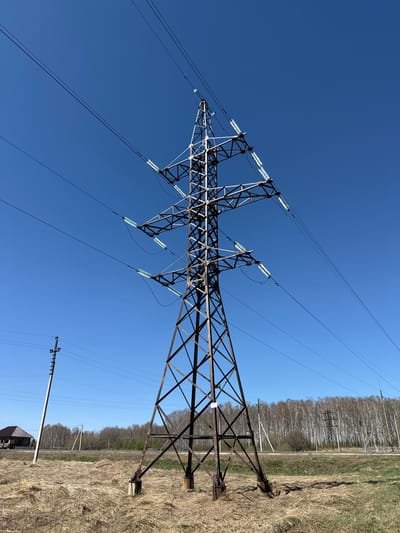 A tall, rusty, metal electrical tower, with numerous wires running from it, stands on a field of dry grass. The tower is positioned against a blue sky, with a few trees visible in the background. The tower is located in a rural area, near a small town or village.  The environment is rural and open, with a dry field and trees visible in the distance.
