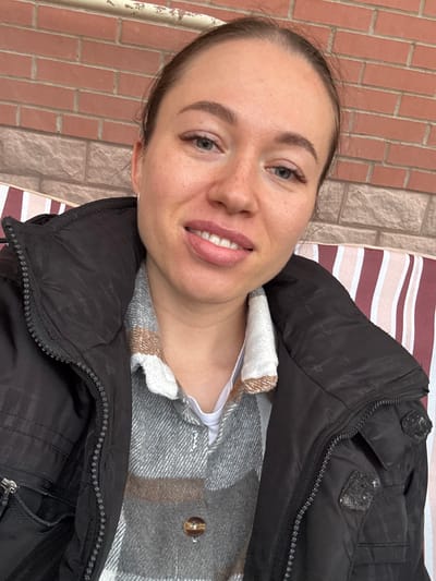 A woman is reclining on a striped surface, likely a lounger or a similar outdoor seating arrangement. She is wearing a grey and white plaid shirt under a black puffer jacket. Her hair is pulled back, and she is smiling slightly, displaying her teeth. In the background, there's a brick wall. The location is Ogryzkovo, Russia.