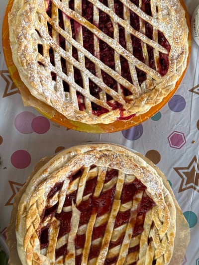 Two berry pies with lattice crusts dusted with powdered sugar sit side-by-side on a table covered with a patterned tablecloth featuring circles, hexagons, and stars. The pie on the left is on an orange plate. The location is not discernible from the image.
