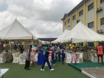 The image shows an outdoor party scene in front of a large yellow building with white windows. The event is taking place in a grassy area, with two large white tents set up to provide shade. There are several people present, including children and adults, and tables are set up with decorations and food. The tables are decorated with a variety of colors, including pink, blue, and yellow. The children are dressed in a variety of attire, with some wearing suits and dresses. Several adults can be seen standing and talking to each other. The image was taken on a cloudy day. The event is being held at a location in Close, Nigeria (latitude: 6.571, longitude: 3.39). There is a blue and white balloon at the bottom left of the image, near a white table. A large window is visible in the upper right corner of the image, featuring what appears to be a metal air conditioner unit. There are several people standing under the tents, and a single person is walking away from the tents towards the left side of the image. This person is wearing a pink top and a blue pair of pants and appears to be leaving the event. A man wearing a pink shirt and black shorts is standing to the right of the image, in front of the yellow building. He appears to be watching the guests at the event. There is a long, thin strip of green artificial grass running parallel to the bottom of the image. The image appears to be a snapshot of a festive and happy event.