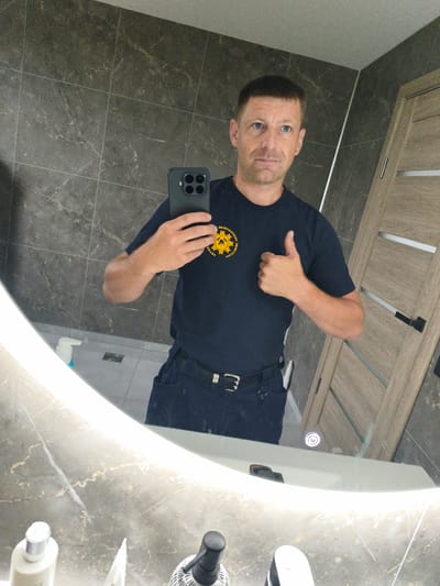 In the city of Kyviškės, Lithuania, a man in a dark blue t-shirt and dark blue pants is taking a mirror selfie in a modern bathroom. The bathroom features dark grey tiled walls with a marble pattern and a wooden door with horizontal glass inserts. The man has short brown hair and blue eyes, and he is wearing a silver necklace and a belt with a silver buckle. On his t-shirt, there is a circular yellow emblem with text that is reversed due to the mirror. He is holding a black smartphone in his left hand and giving a thumbs-up gesture with his right hand. A large, illuminated mirror with a marble-effect frame dominates the foreground, reflecting the man and parts of the bathroom. Various toiletries are visible on the counter below the mirror.