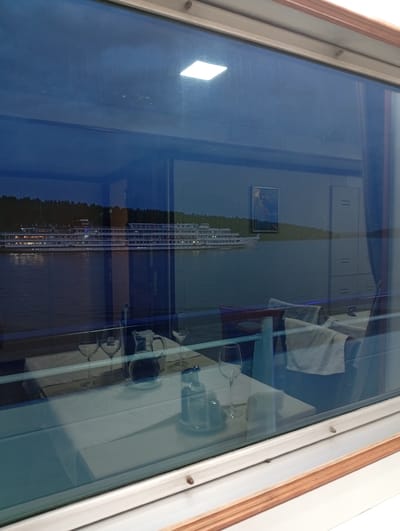 A view through a window shows a dining table set for service. On the table, there are wine glasses, a water pitcher, and other tableware on a white tablecloth. The window reflects the outside view: a multi-deck river cruise ship is passing, with a backdrop of trees on the shore. Inside, a partially visible chair and other dining room furnishings can be seen. The image was taken from inside a building, likely a restaurant or dining area, that is situated somewhere in Troitskiy Posad, Russia, as indicated by the identified objects and location.