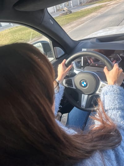 An overhead, slightly tilted shot captures a person in the driver's seat of a BMW car, likely in Saurieši, Latvia. The driver, with long brown hair flowing over their shoulder, wears a light blue fuzzy sweater and blue jeans. Their hands are on the steering wheel, with the left hand at the top and the right hand appearing to adjust controls on the lower spoke. A silver watch is visible on the left wrist.
The car's interior is modern, featuring a BMW M Sport steering wheel with the BMW logo prominently displayed. The digital dashboard is partially visible, showing "Power % 100", "D" (for Drive), "49" (possibly speed or remaining range), and "COMFORT" mode selected. Other visible text on the steering wheel includes "RESUME CANCEL SET". On the A-pillar, the word "AIRBAG" is visible. A white strap or belt with "LOEWE" printed on it is seen near the driver's lap.
Through the front windshield, a paved road stretches ahead with a grassy area and some distant structures to the left. The scene suggests bright daylight, with clear skies and good visibility. The time displayed on the dashboard is "10:56". The person appears to be driving or preparing to drive.