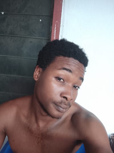 This is a close-up, eye-level selfie of a young Black man, likely in his late teens or early twenties, from the chest up. He is facing the camera with a neutral expression, looking directly at the viewer. He has dark, curly hair and a light beard and mustache. His skin is brown.

The background is divided into two distinct areas. To the left is a dark, textured surface that appears to be a corrugated metal or plastic wall, possibly part of a building's exterior or a makeshift structure. To the right, the background is a bright, white, textured surface, suggesting a wall or plastered surface. A thin, reddish-brown vertical stripe runs down the center between these two surfaces.

The lighting suggests it is daytime, with natural light illuminating the man's face from the right. The overall setting appears to be outdoors or in an area with natural light, possibly in a less developed or residential area given the background textures. There are no visible objects other than the man and the background elements. The man is shirtless, and his shoulders and upper chest are visible. The image conveys a sense of casualness and directness.